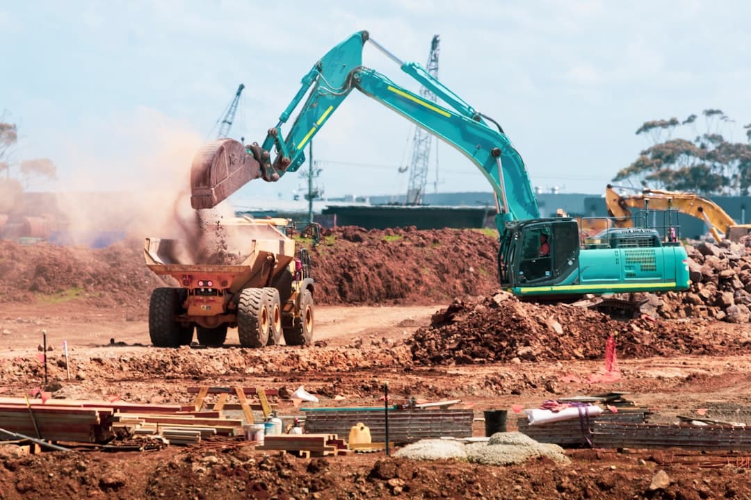 A teal excavator dumping soil into a yellow dump truck at a construction site